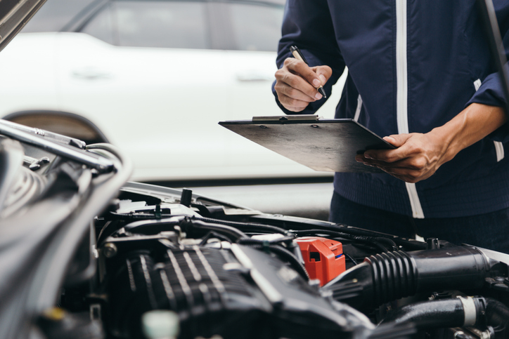 Exposed car engine and mechanic in a navy jacket referring to a checklist during a car service book.